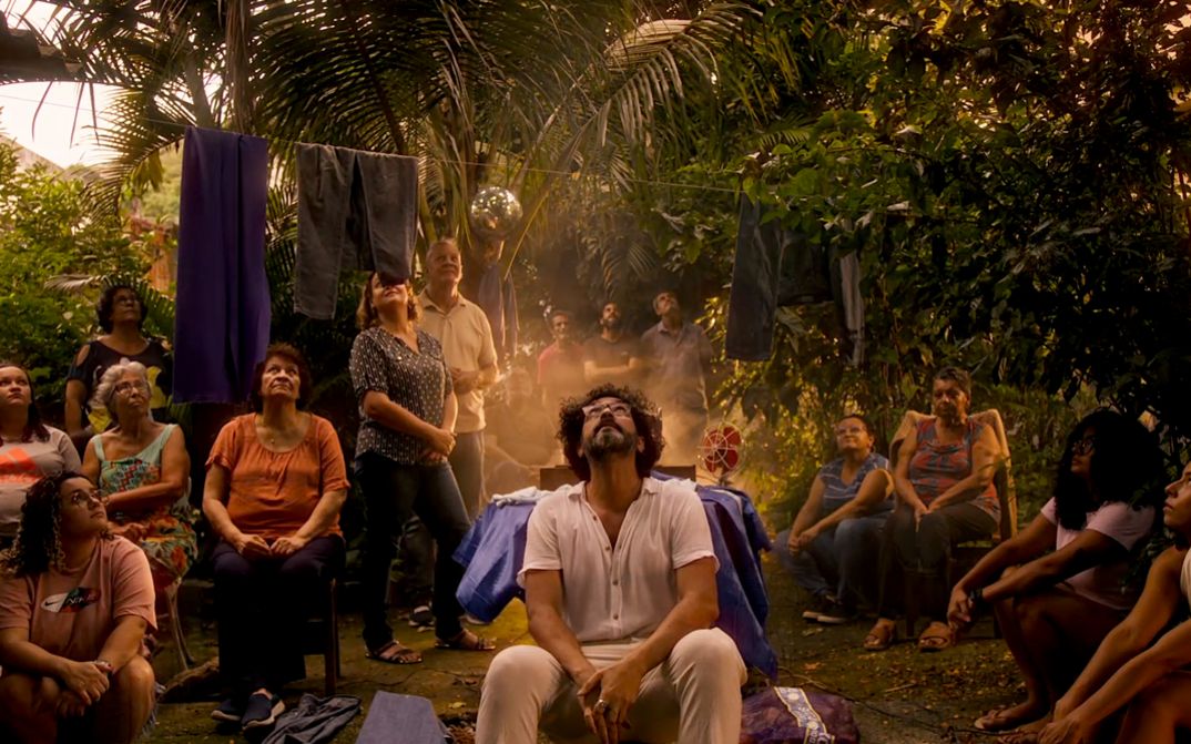 Filmstill aus dem Film „Zizi (ou oração da jaca fabulosa)“ von Felipe M. Bragança. Im Innenhof eines Hauses mit üppiger Vegetation blickt eine generationenübergreifende Gruppe zum Himmel. Der Protagonist sitzt in der Mitte der Gruppe, während ein goldenes, ätherisches Licht vom Boden aufsteigt und die Szene in eine übernatürliche Aura taucht.
