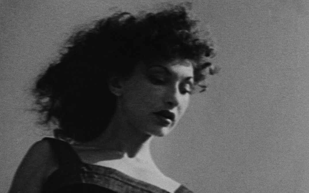 Filmstill aus AT LAND: Schwarz-weiß-Aufnahme einer Frau mit dunklen Locken, in leichter Untersich aufgenommen.