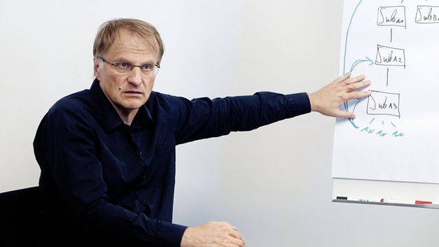 Still from the film "For the Many – The Vienna Chamber of Labour" by Constantin Wulff: A man in a seminar room is sitting and pointing to a flip chart with notes on it