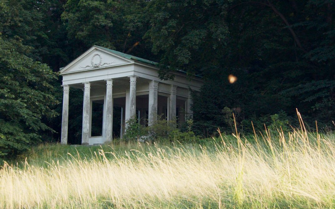 Filmstill aus „De Facto" von Selma Doborac. Ein Gebäude mit Säulen am Rande eines Waldes.