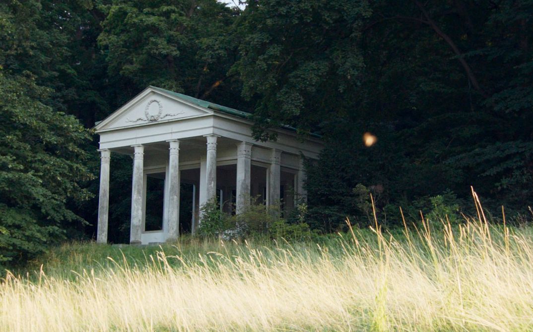 Filmstill aus „De Facto" von Selma Doborac. Ein Gebäude mit Säulen am Rande eines Waldes.