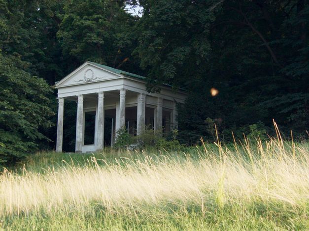 Filmstill aus „De Facto" von Selma Doborac. Ein Gebäude mit Säulen am Rande eines Waldes.