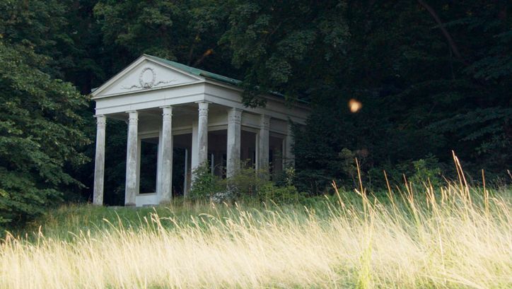 Filmstill aus „De Facto" von Selma Doborac. Ein Gebäude mit Säulen am Rande eines Waldes.