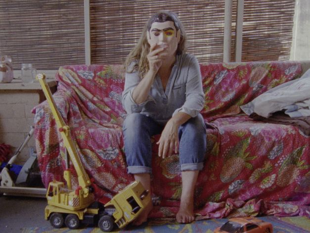 Film still from EVIDENCE by Lee Anne Schmitt. The image shows a person sitting on a sofa covered with a pink cloth and holding a mask in front of their face. On the floor is a toy crane.