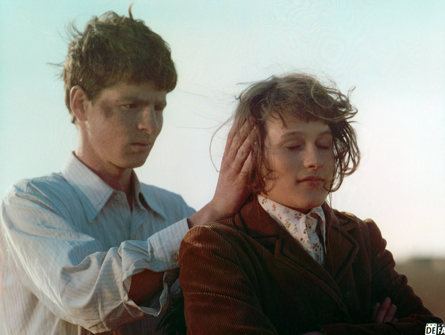 Filmstill aus ROMANZE MIT AMÉLIE: Ein junger Mann steht hinter einer jungen Frau und legt seine Hände an ihre Ohren. 