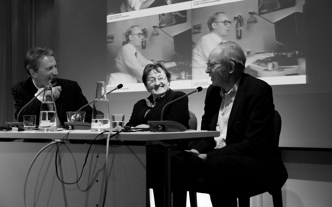 Erika und Ulrich Gregor mit Ralph Eue Ulrich und Erika Gregor sitzen mit Ralph Eue auf einer Bühne. Hinter ihnen ist auf die Leinwand ein Foto von Ulrich Gregor in jüngeren Jahren projiziert.