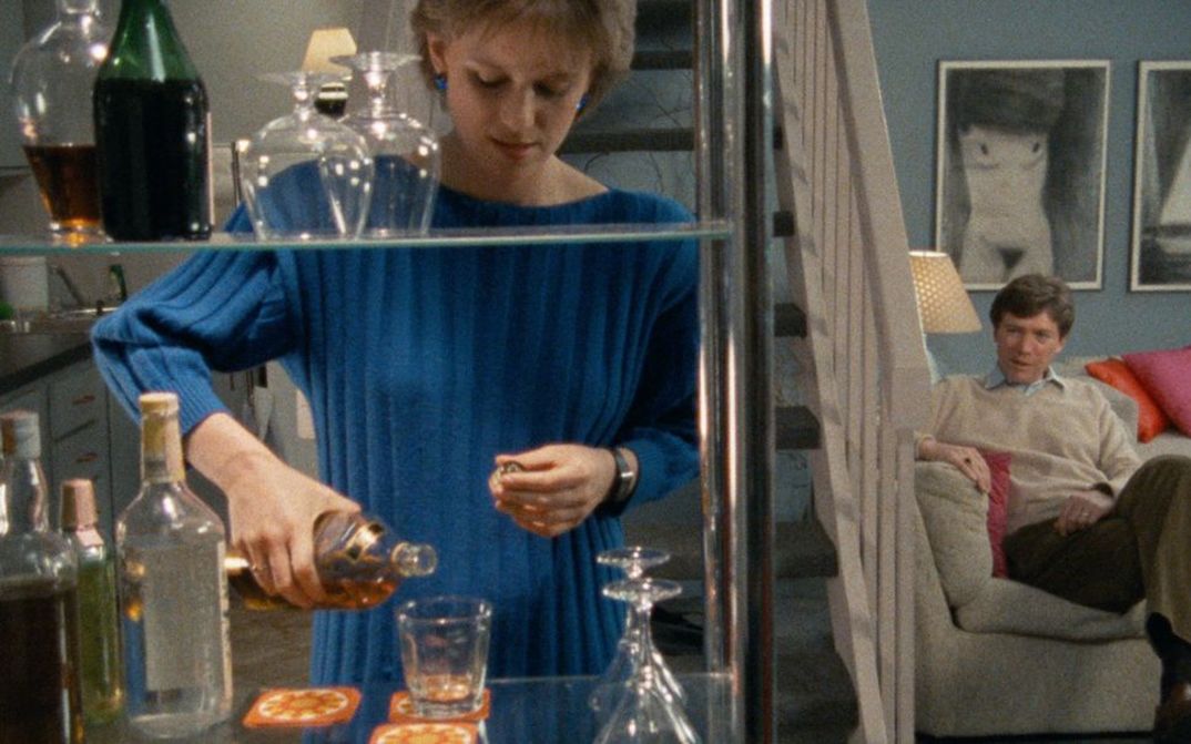 Filmstill aus WORKING GIRLS. Eine elegant gekleidete Frau schenkt in einer Wohnung einen Drink ein, im Hintergrund sitzt ein Mann auf einem Sofa.