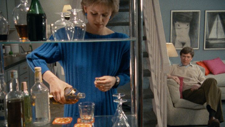 Filmstill aus WORKING GIRLS. Eine elegant gekleidete Frau schenkt in einer Wohnung einen Drink ein, im Hintergrund sitzt ein Mann auf einem Sofa.