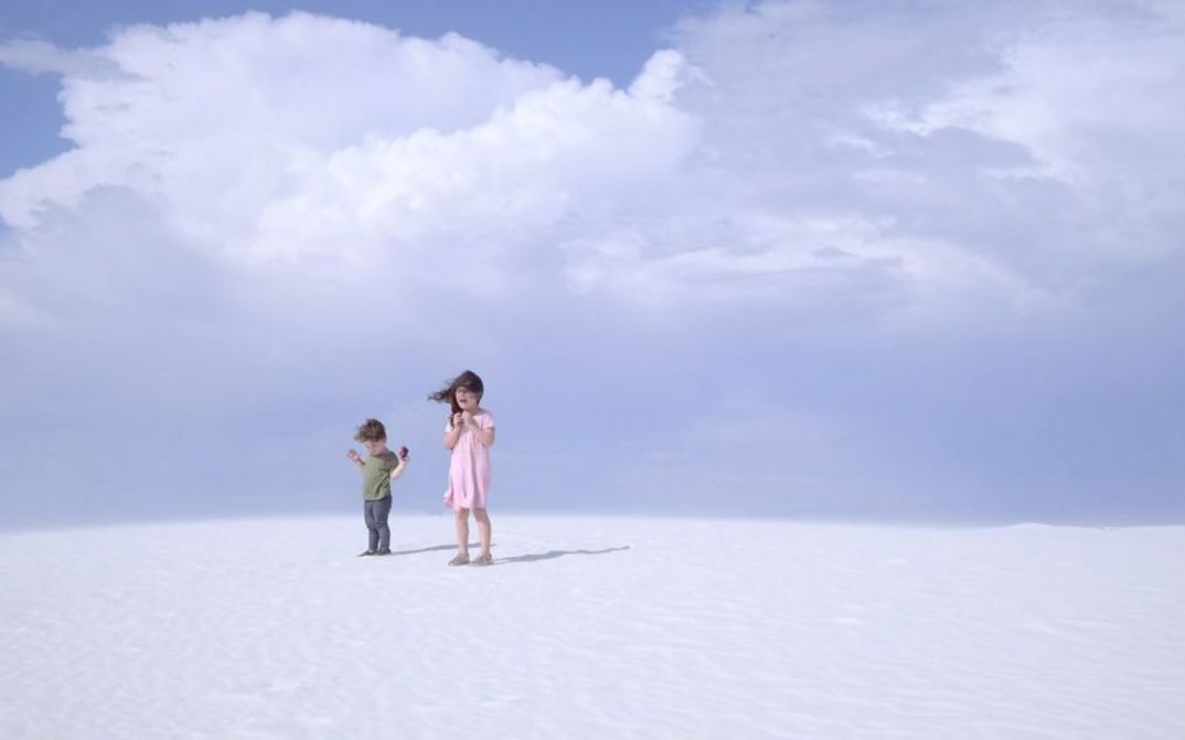 Still from the film "Nuclear Family" by Erin Wilkerson and Travis Wilkerson. Two children stand on a sand dune in front of a blue sky.