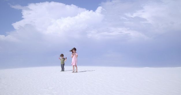 Still from the film "Nuclear Family" by Erin Wilkerson and Travis Wilkerson. Two children stand on a sand dune in front of a blue sky.