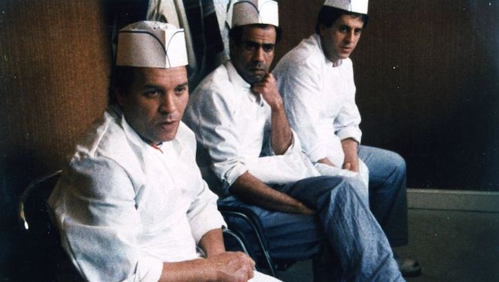 Film still from COÛTE QUE COÛTE: Three men in white cooking clothes sit next to each other.