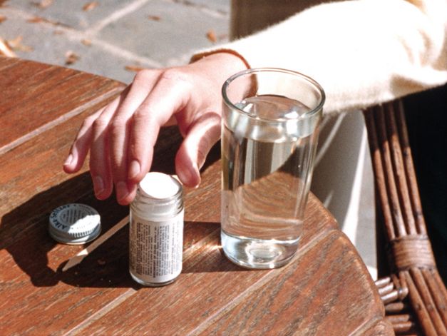 Still from the film "Home When You Return" by Carl Elsaesser. A hand reaches for a medicine that is on a table next to a glass of water.