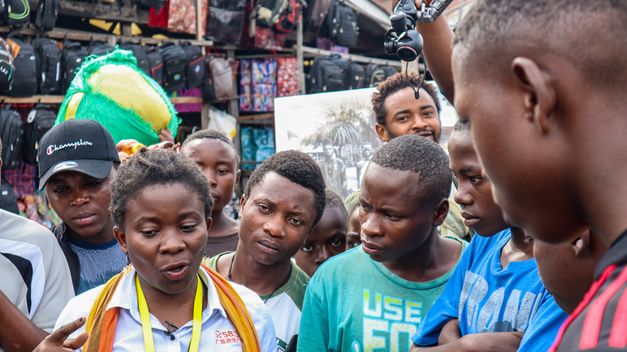 Still from the Film "Kumbuka" by Petna Ndaliko Katondolo. A group of people stand close around a woman and listen to her speak.