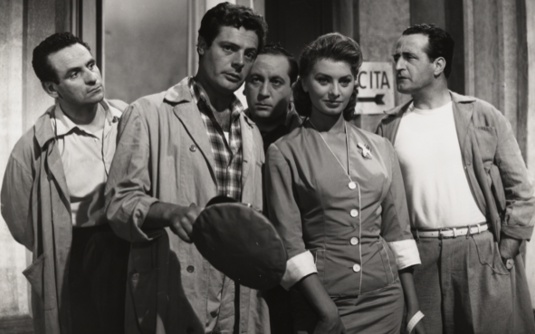Film still from PECCATO CHE SIA UNA CANAGLIA: Marcello Mastroianni in work clothes and Sophia Loren are standing next to each other, behind them three men, all of whom are looking at Mastroianni.