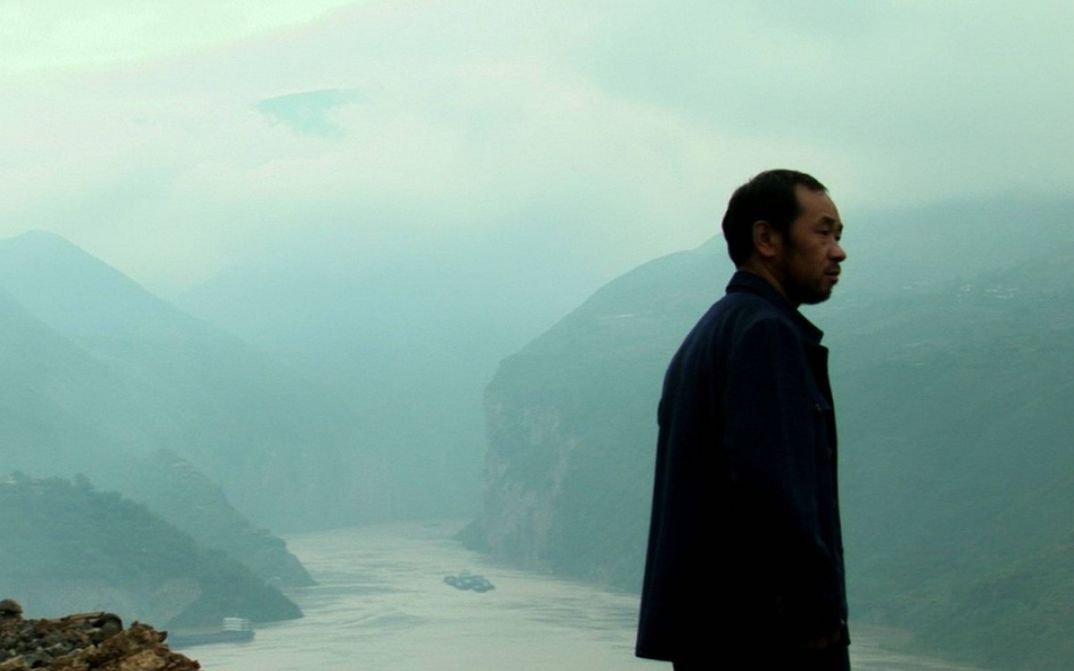 Filmstill from STILL LIFE: A man stands on high ground and looks to the right. A river and mountains below him. It is foggy.