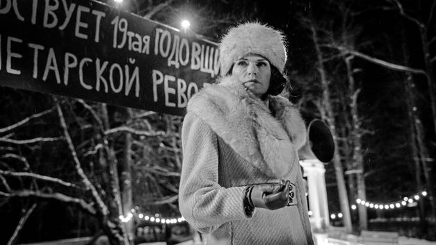 Film still from "Marijas klusums" by Dāvis Sīmanis. It shows a black-and-white image of a woman in the dark wearing a coat, fur scarf, fur hat and leather gloves. 