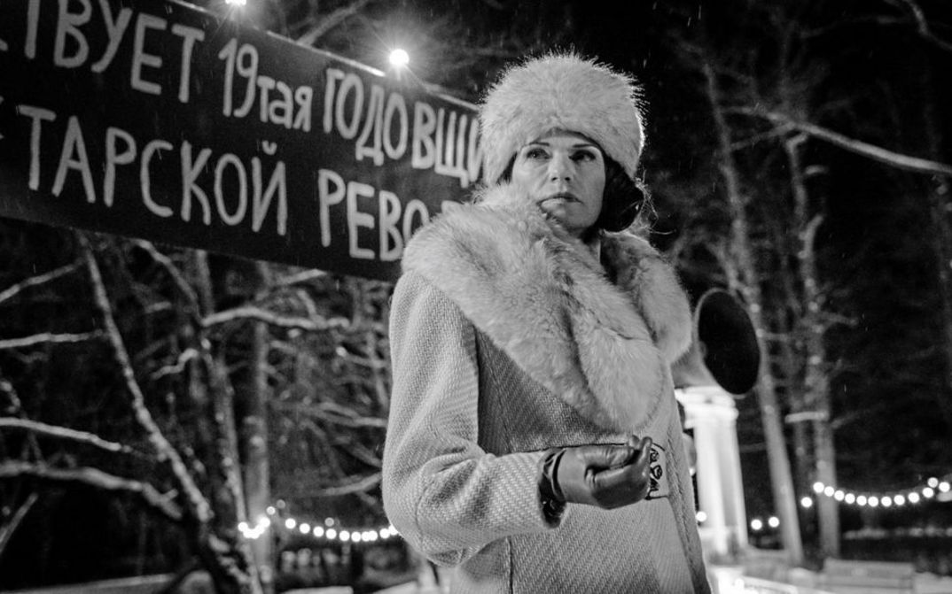 Filmstill aus "Marijas klusums" von Dāvis Sīmanis. Zu sehen ist eine Schwarz-Weiß-Aufnahme einer Frau im Dunkeln mit Mantel, Pelzschal, Pelzmütze und Lederhandschuhen. 