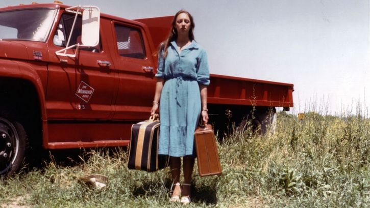 Filmstill aus EMPTY SUITCASES: Eine Frau steht mit zwei Koffern in beiden Händen vor einem roten Pick-up-Truck auf einer Wiese.