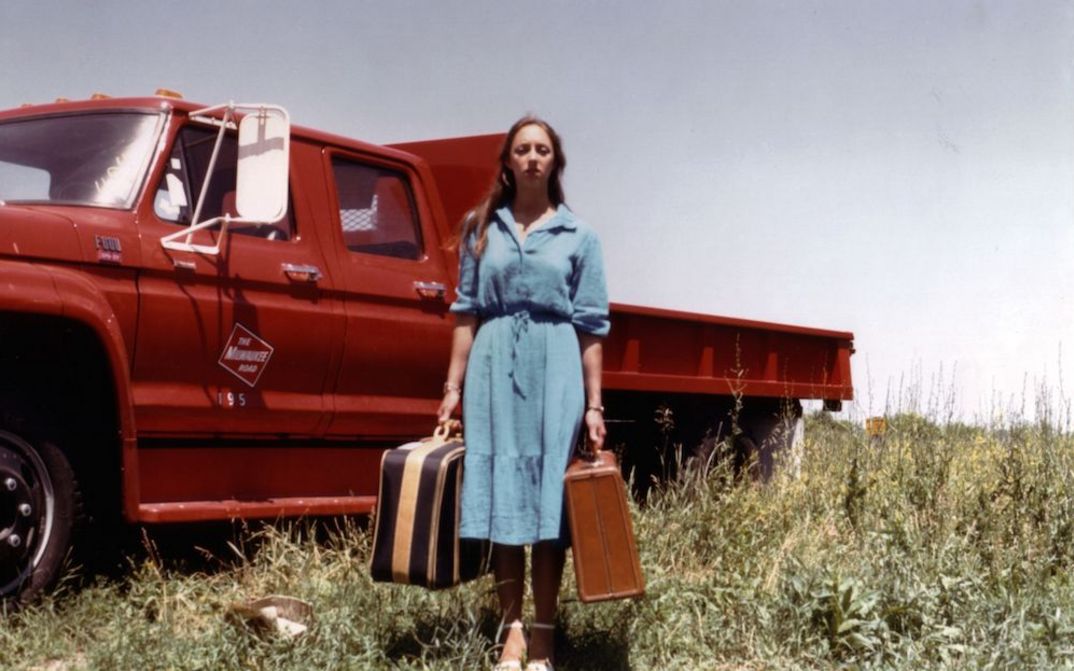 Filmstill aus EMPTY SUITCASES: Eine Frau steht mit zwei Koffern in beiden Händen vor einem roten Pick-up-Truck auf einer Wiese.