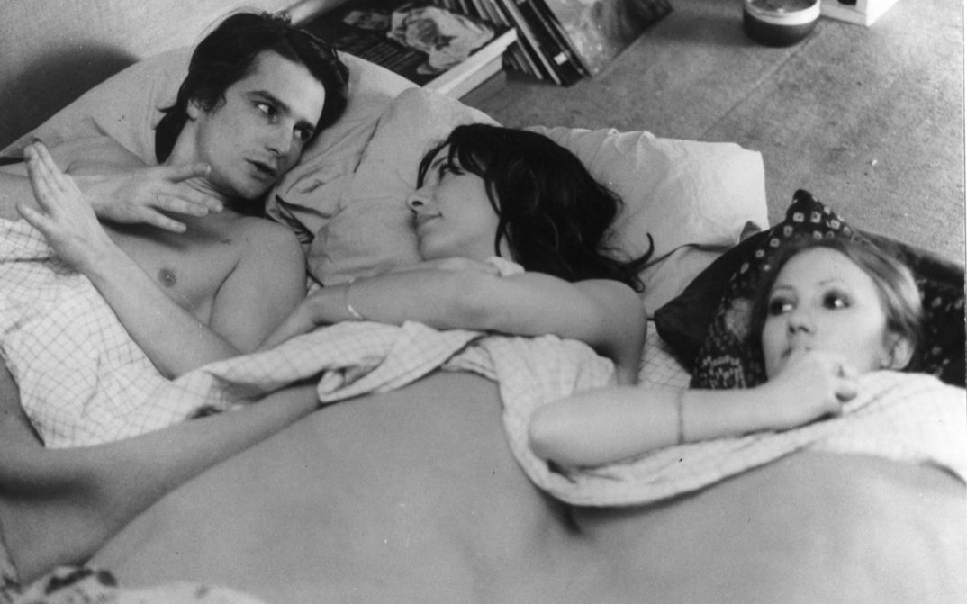 Filmstill aus LA MAMAN ET LA PUTAIN: Ein Mann und zwei Frauen liegen nebeneinander in einem Bett und unterhalten sich.