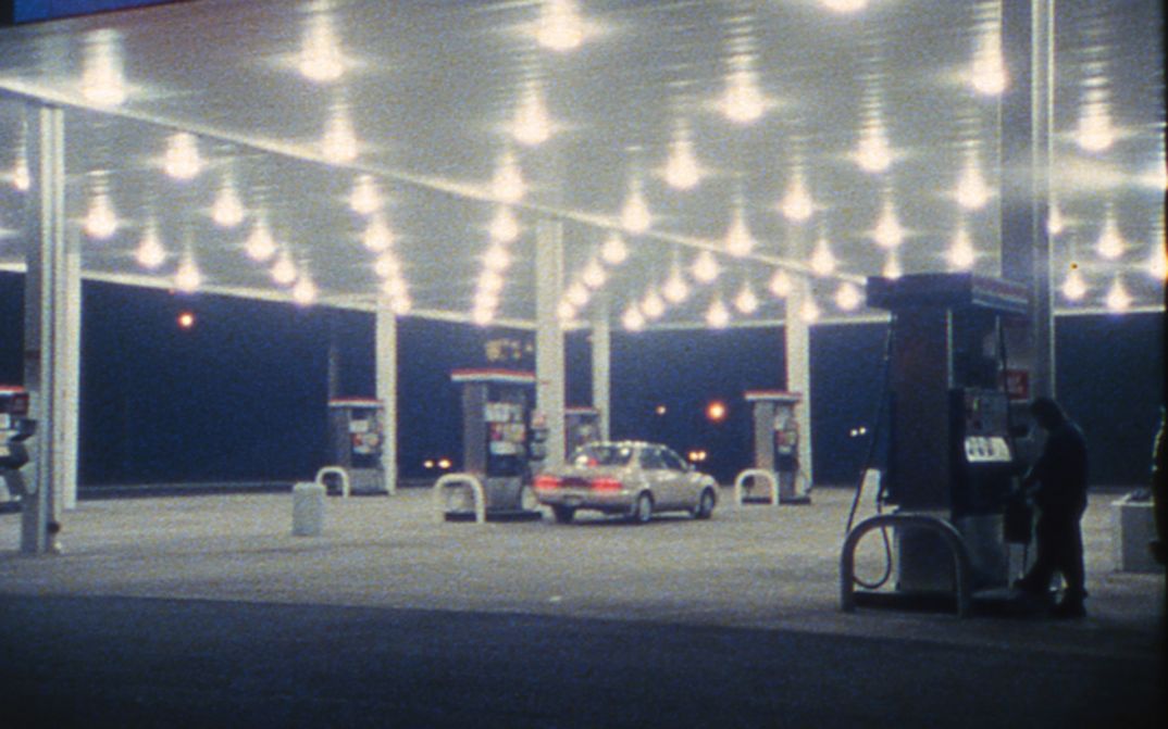 Filmstill aus SOMETHING MORE THEN NIGHT: Eine Tankstelle im Dunkeln. Ein Auto steht an der Zapfsäule in der Mitte.