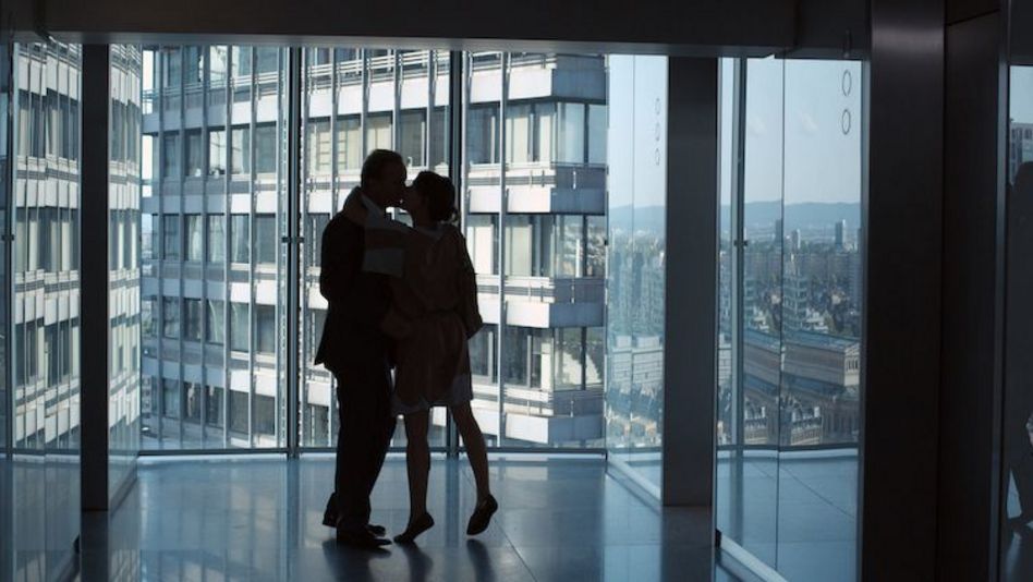 Film still from UNTER DIR DIE STADT: In an empty room in a high-rise building with glass facades, two people are kissing. Outside, more high-rise buildings can be seen.