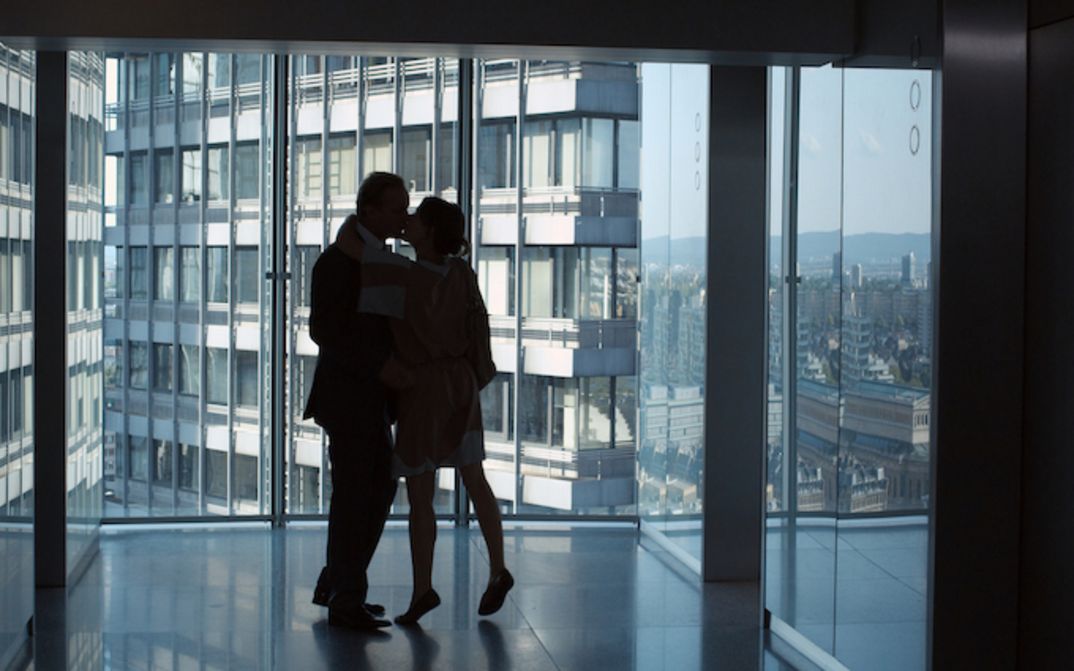Filmstill aus UNTER DIR DIE STADT: In einem leeren Raum in einem Hochhaus mit Glasfassaden küssen sich zwei Personen. Draußen sind weitere Hochhäuser zu sehen.