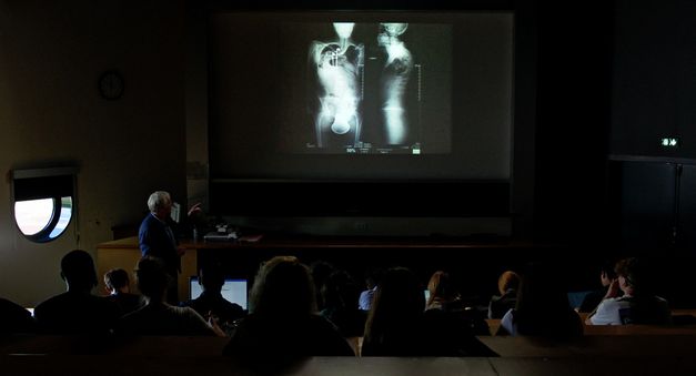 Filmstill aus "Europe" von Philipp Scheffner. Ein Professor und Studierende in einem abgedunkelten Vorlesungssaal. Auf der Leinwand ist ein vergrößertes Röntgenbild eines Menschen zu sehen. 