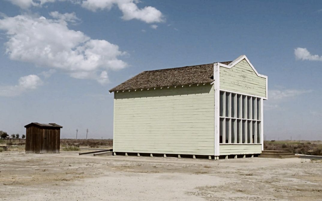 Filmstill aus dem Film „Allensworth" von James Benning. Ein hellgelbes Holzhaus vor einer kargen Landschaft und blauem Himmel.