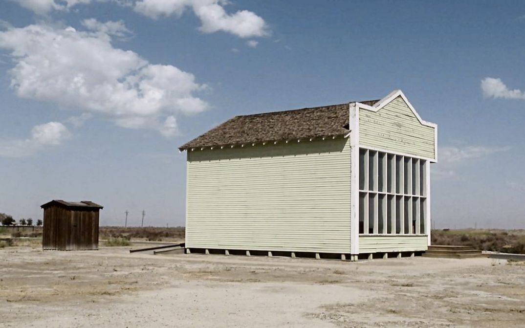 Filmstill from "Allensworth" by James Benning. A light yellow wooden house against a barren landscape and blue sky.