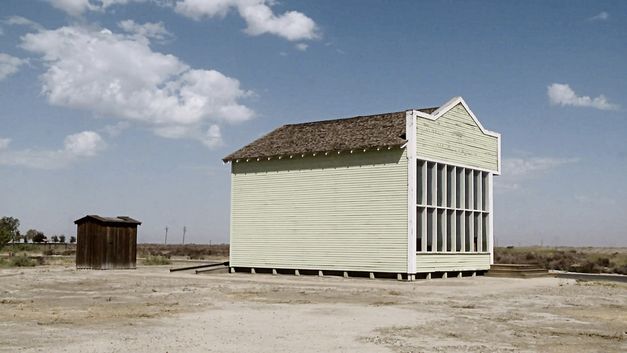 Filmstill from "Allensworth" by James Benning. A light yellow wooden house against a barren landscape and blue sky.