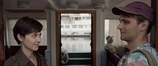 Film still from "Redaktsiya" by Roman Bondarchuk. It shows a woman with a short haircut and a man with a cap standing opposite each other in a room and looking at each other. A glass door can be seen in the background and another person is sitting at a table. 