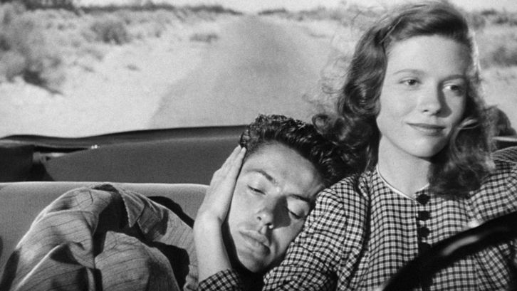 Filmstill aus THEY LIVE BY NIGHT: Eine junge Frau und ein junger Mann fahren in einem offenen Auto durch eine weite, leere Landschaft. Sie sitzt am Steuer und hält seinen Kopf, den er an ihre Schulter gelehnt hat. 
