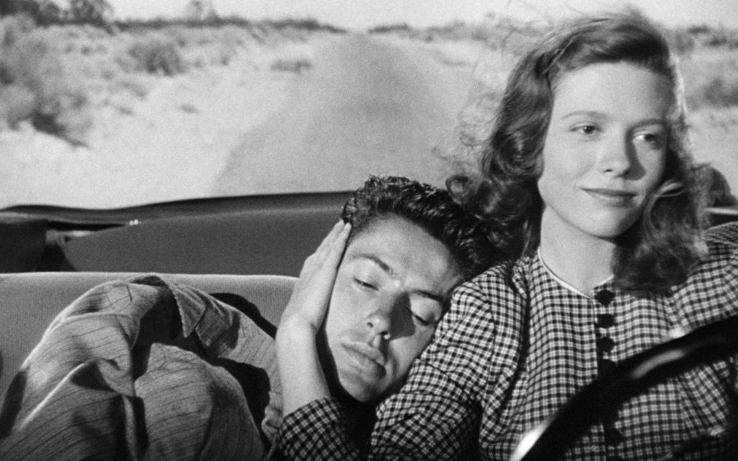 Filmstill aus THEY LIVE BY NIGHT: Eine junge Frau und ein junger Mann fahren in einem offenen Auto durch eine weite, leere Landschaft. Sie sitzt am Steuer und hält seinen Kopf, den er an ihre Schulter gelehnt hat.