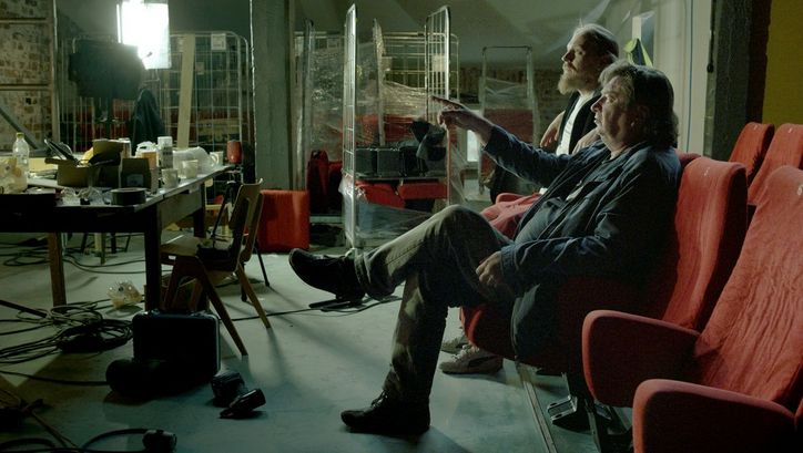 Film still from CINÉMA LAIKA: Two men are sitting on movie theater chairs in a somewhat chaotically cluttered room. One of them is the director Aki Kaurismäki.