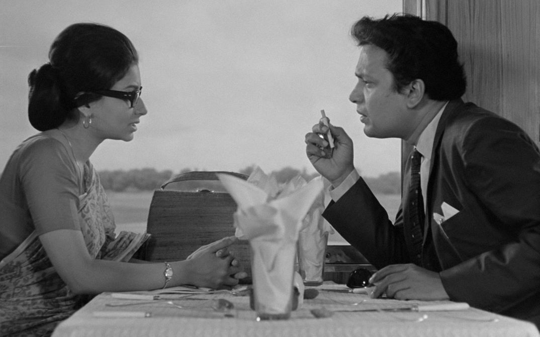 Filmstill aus NAYAK: Ein Mann und eine Faru sitzen an einem Restauranttisch am Fenster. Der Mann wirkt impulsiv, er hält eine Zigarette in der Hand.