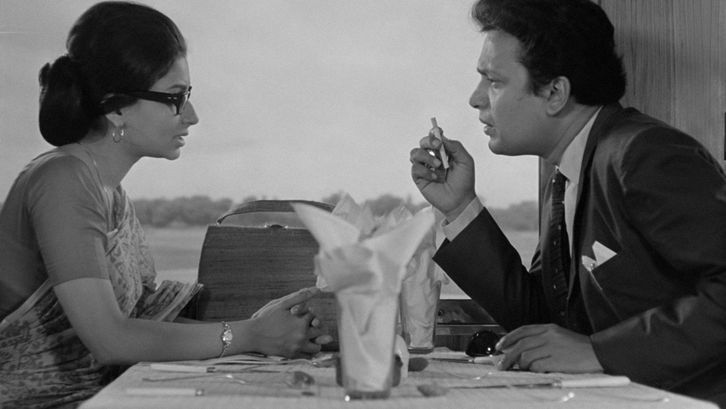 Filmstill from NAYAK: A man and a woman are sitting at a restaurant table by the window. The man seems impulsive, he is holding a cigarette in his hand.