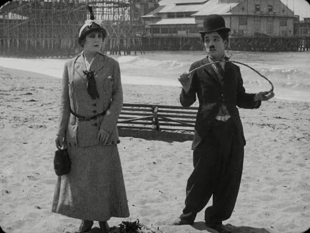 Filmstill von BY THE SEA: Charlie Chaplin und eine Frau stehen nebeneinander am Strand.