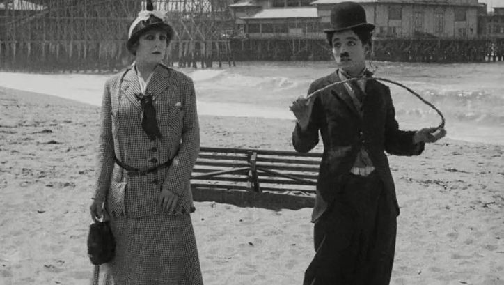 Filmstill von BY THE SEA: Charlie Chaplin und eine Frau stehen nebeneinander am Strand.