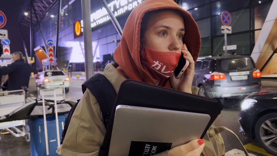 Filmstill aus MY UNDESIRABLE FRIENS: Eine junge Frau steht zwischen Autos vor einem Flughafen. Sie trägt eine Jacke mit Kapuze, hat einen Laptop in der Hand und hält ihr Telefon ans Ohr.