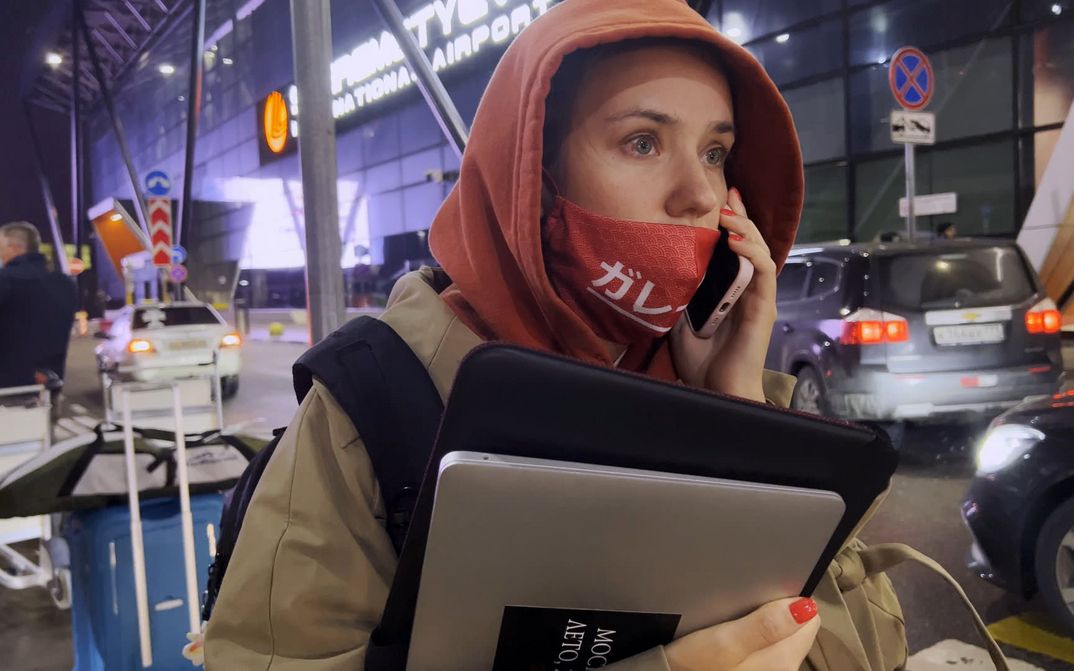 Filmstill aus MY UNDESIRABLE FRIENS: Eine junge Frau steht zwischen Autos vor einem Flughafen. Sie trägt eine Jacke mit Kapuze, hat einen Laptop in der Hand und hält ihr Telefon ans Ohr.
