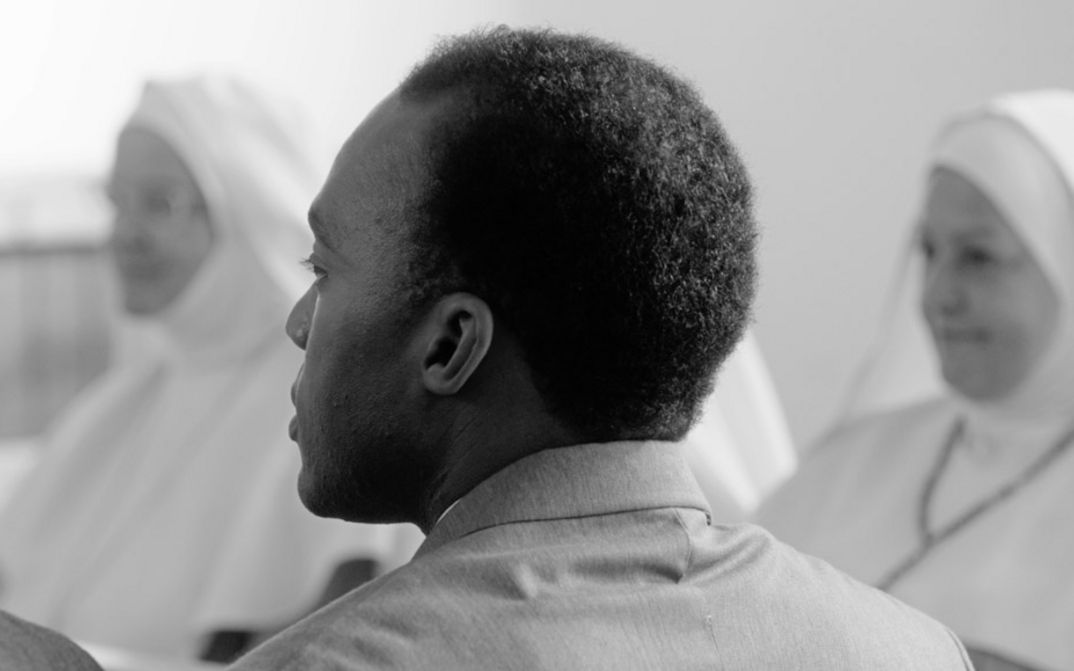 Filmstill aus "Chroniques fidèles survenues au siècle dernier à l’hôpital psychiatrique Blida-Joinville, au temps où le Docteur Frantz Fanon était chef de la cinquième division entre 1953 et 1956" von Abdenour Zahzah. Zu sehen ist eine schwarz-weiße Nahaufnahme eines Hinterkopfes einer Person mit einer Kurzhaarfrisur. Im Hintergrund sind ganz in Weiß gekleidete Menschen zu sehen.  