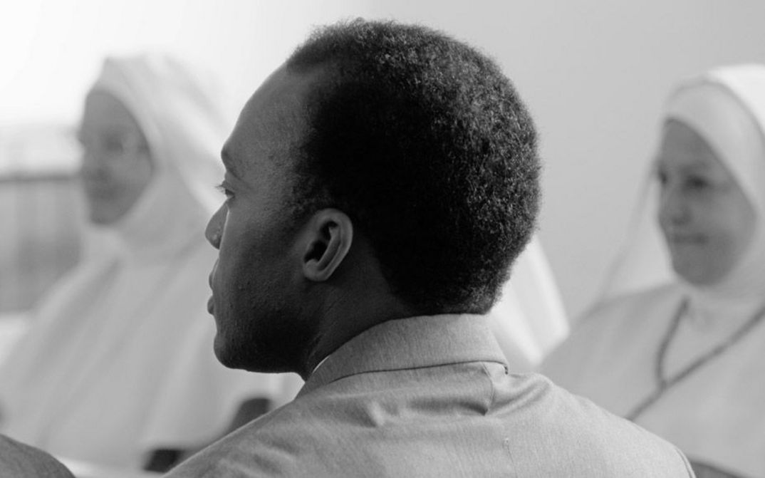 Filmstill aus "Chroniques fidèles survenues au siècle dernier à l’hôpital psychiatrique Blida-Joinville, au temps où le Docteur Frantz Fanon était chef de la cinquième division entre 1953 et 1956" von Abdenour Zahzah. Zu sehen ist eine schwarz-weiße Nahaufnahme eines Hinterkopfes einer Person mit einer Kurzhaarfrisur. Im Hintergrund sind ganz in Weiß gekleidete Menschen zu sehen.