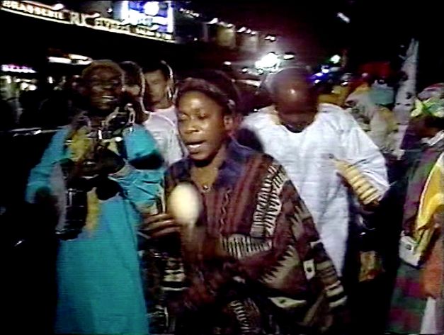 Still from the film "Moune Ô" by Maxime Jean-Baptiste. People walk through a crowded street with musical instruments in their hands.