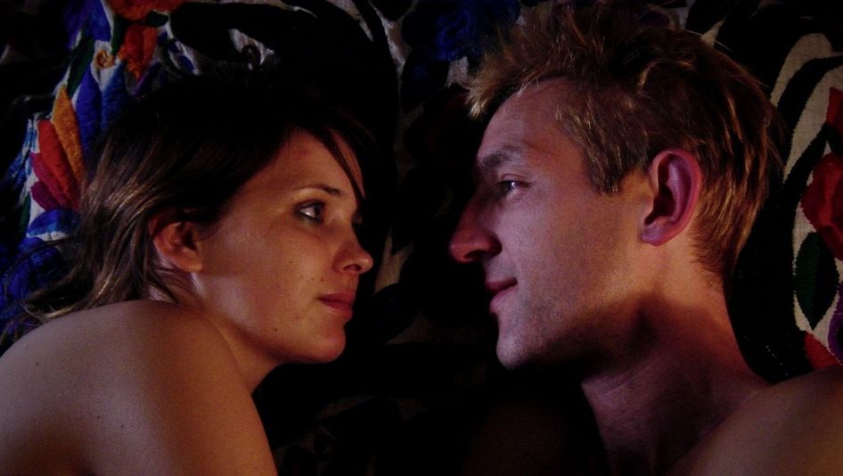 Film still from PIGS WILL FLY: A woman and a man are lying naked side by side in bed, looking into each other's eyes.
