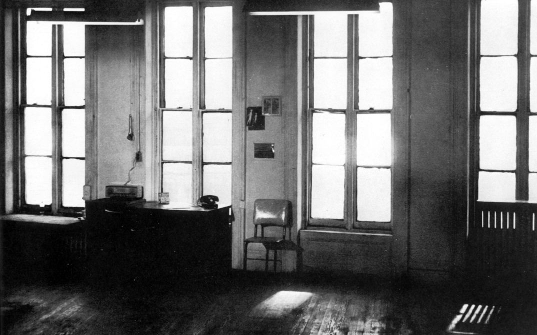 Filmstill aus WAVELENGHT: Man blickt in einem großen Zimmer auf die Fensterfront mit vier Fenstern. Davor stehen ein Schreibtisch und ein Stuhl.