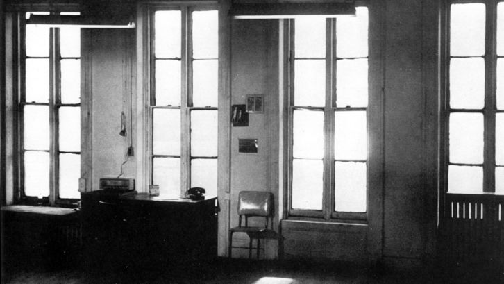 Filmstill aus WAVELENGHT: Man blickt in einem großen Zimmer auf die Fensterfront mit vier Fenstern. Davor stehen ein Schreibtisch und ein Stuhl.