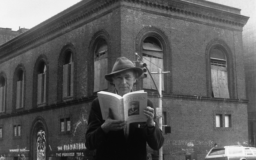 Black and white photograph of Jonas Mekas standing on a street corner in New York, in his hands a book about Maya Deren. In the background is the Anthology Film Archives building.