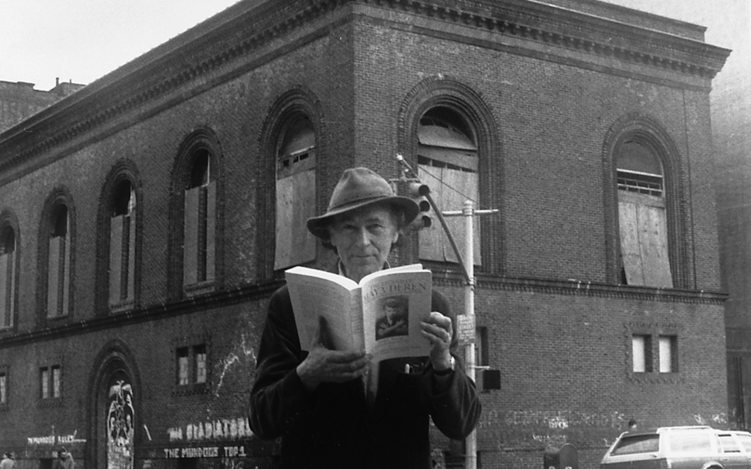 Schwarz-weiß-Fotografie von Jonas Mekas, der in New York an einer Straßenecke steht, in den Händen ein Buch über Maya Deren. Im Hintergrund ist das Gebäude der Anthology Film Archives.
