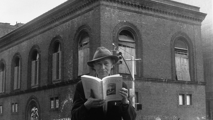 Black and white photograph of Jonas Mekas standing on a street corner in New York, in his hands a book about Maya Deren. In the background is the Anthology Film Archives building.